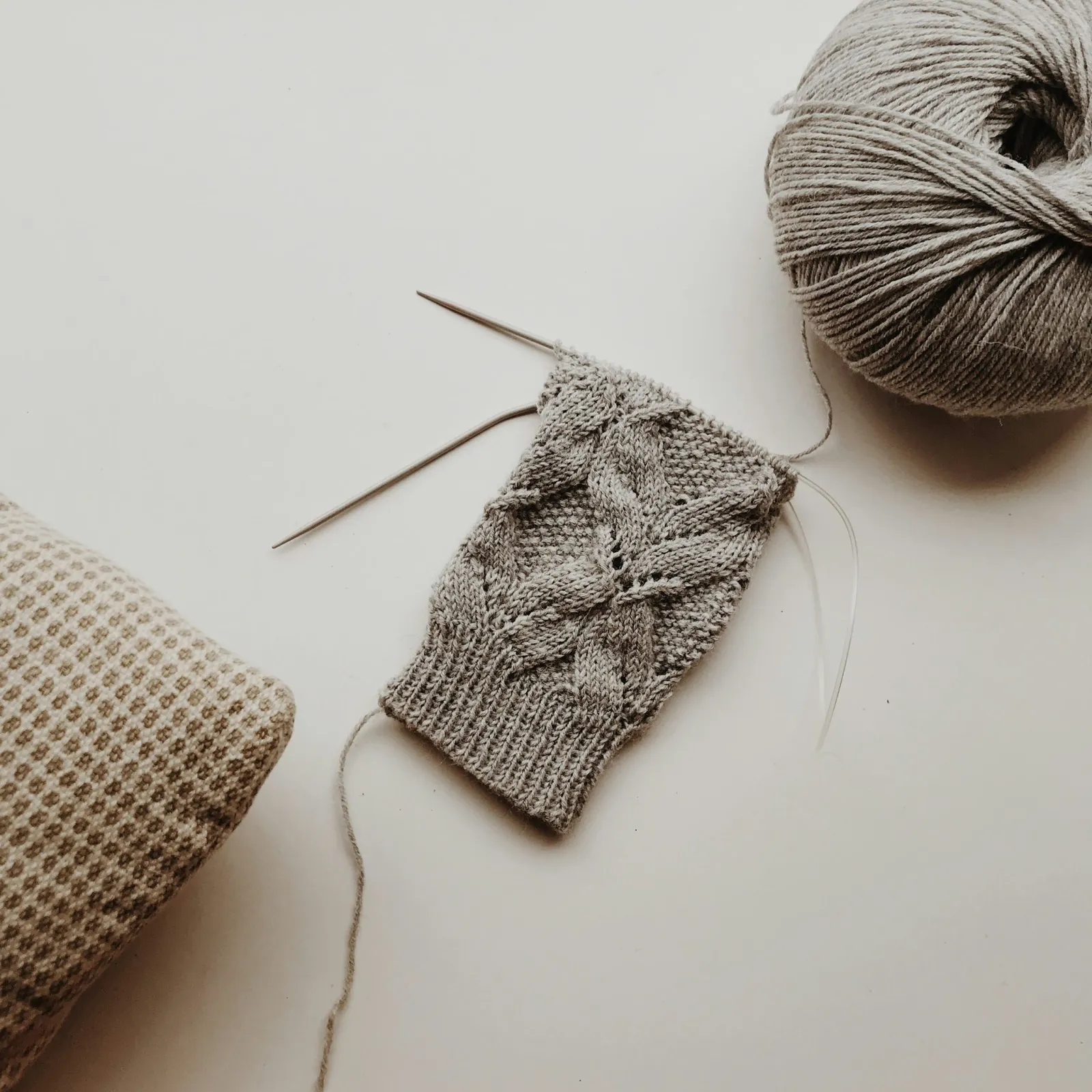 A grey cable knit on needles beside a skein of yarn