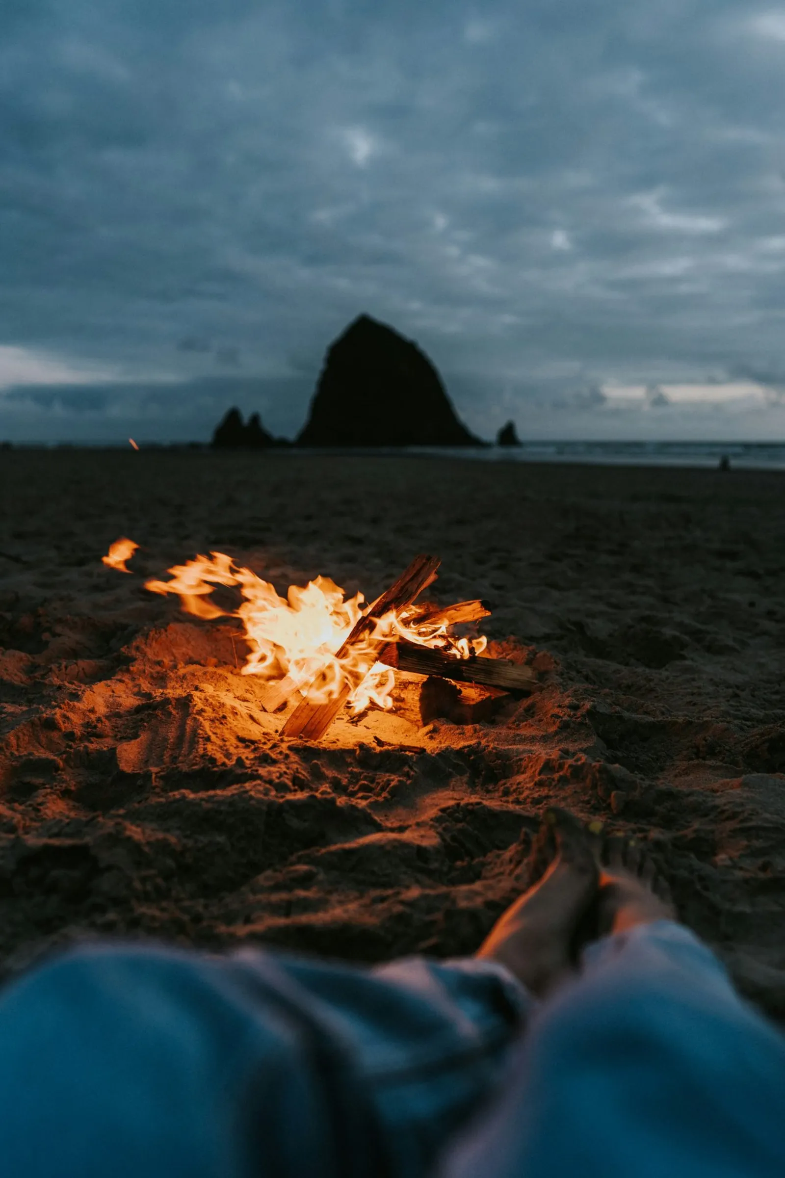 A campfire on a beach at dusk with a person lying nearby